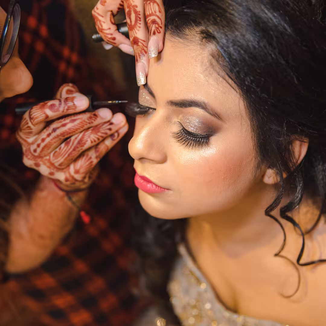 Close-up of a young woman with closed eyes getting eyeliner applied by a makeup artist with henna-decorated hands, wearing a sparkly outfit for a special occasion.