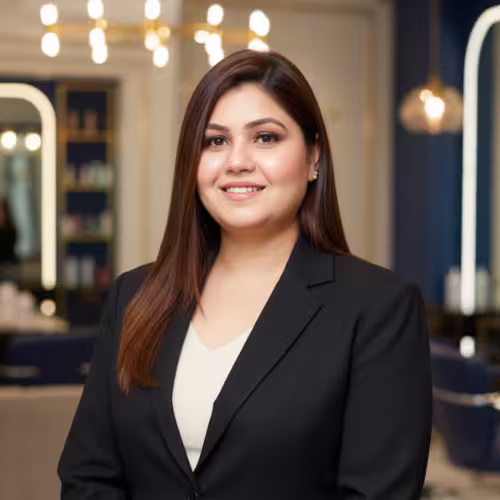 Ruchika Rana smiling confidently in a black blazer, standing in a modern luxury beauty salon with soft blurred background, inviting viewers to chat with her.