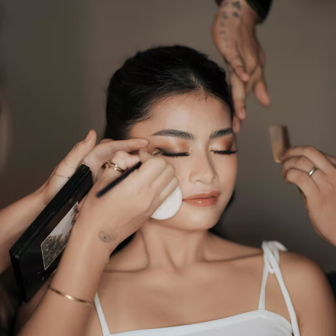 Young woman with eyes closed receiving professional makeup application by two makeup artists in a calm, softly lit beauty studio.