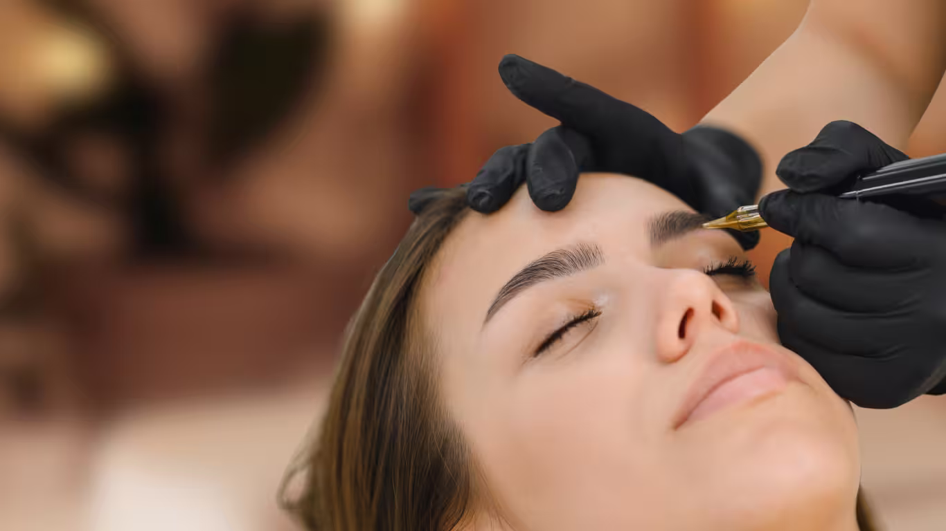 Beautician carefully shaping client’s eyebrows with precision tool during permanent makeup session in a modern salon.